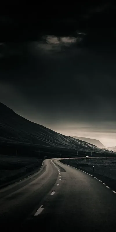 An empty two-lane road bends between dark hills, heavy cloud pressing down and a thin strip of pale light left along the horizon.