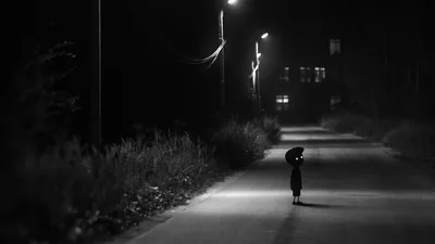 A single streetlamp lights the patch of lane where a figure pauses under an umbrella, weeds crowding the left verge and windows glowing far behind.