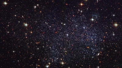 Thousands of resolved stars fill the frame edge to edge in a deep-field view, gold and blue giants flaring among faint red dust.