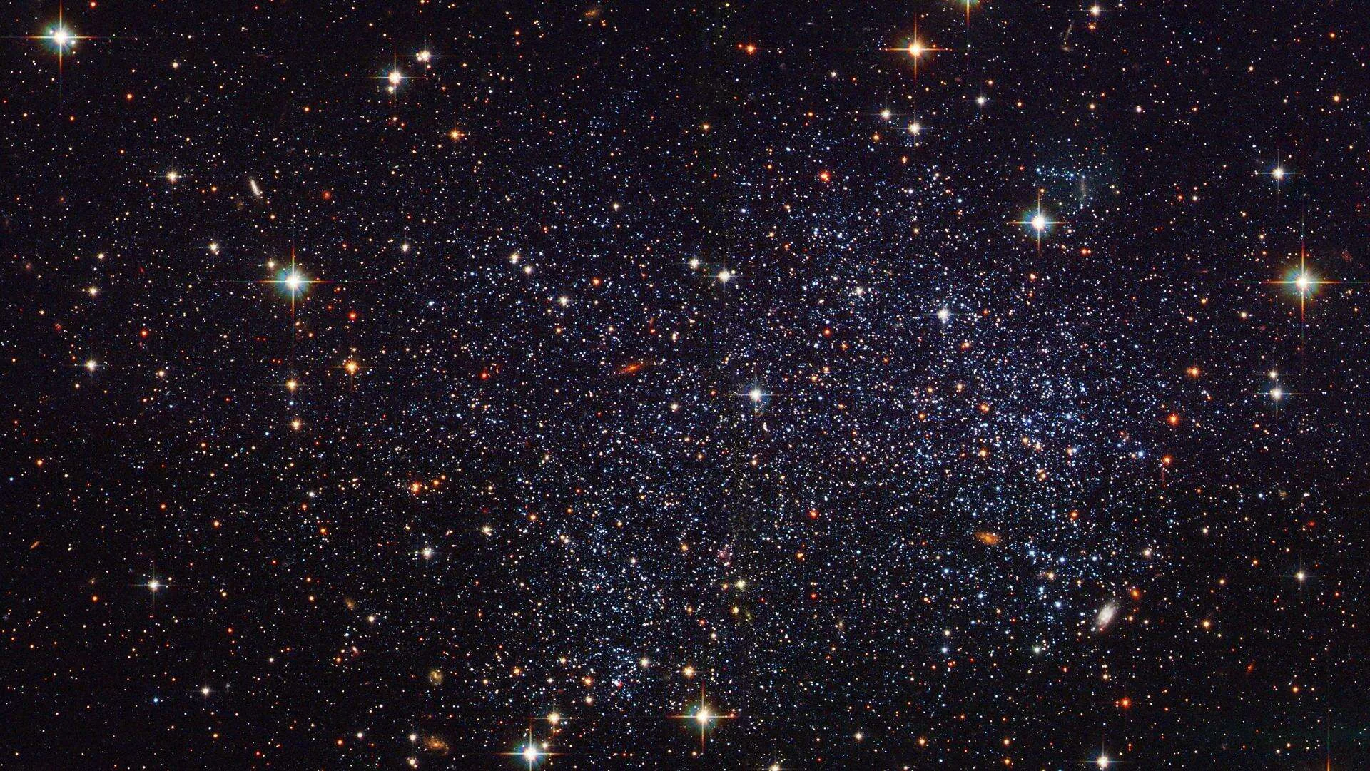 Thousands of resolved stars fill the frame edge to edge in a deep-field view, gold and blue giants flaring among faint red dust.