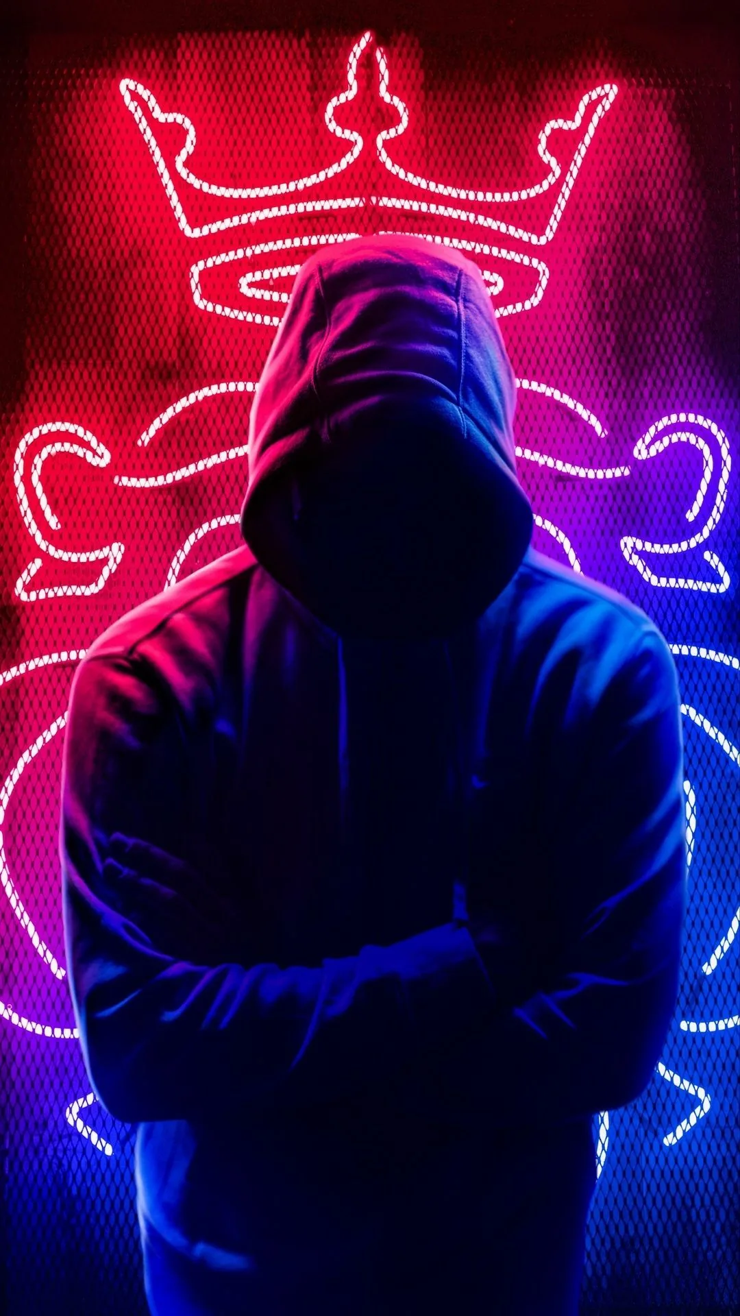 Arms crossed and face hidden, he stands against a wall sign of red and blue tubing that outlines a crown and lettering.