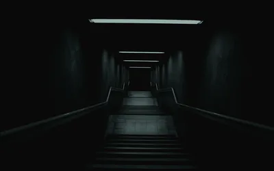 A staircase climbs toward a lit doorway, a single strip light overhead and the walls left almost black.