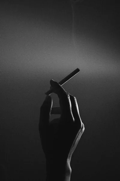 A hand lifts a cigarette into a grey gradient, thin smoke rising past the top edge while the arm stays in shadow.