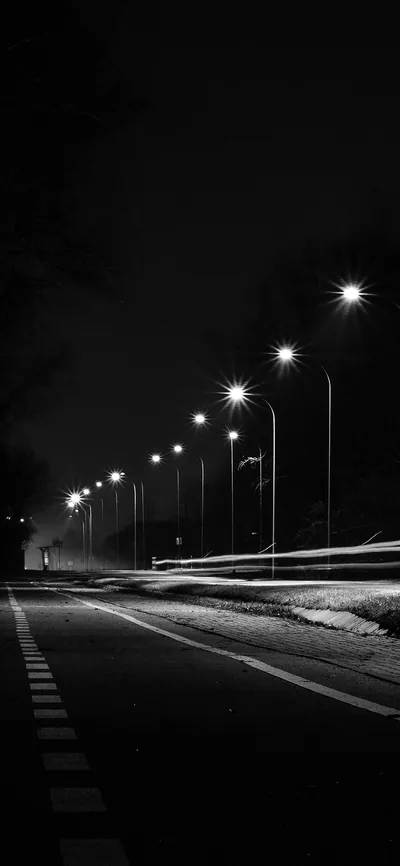 A row of street lights recedes down an empty road at night in black and white, headlight trails streaking the tarmac at mid-frame.