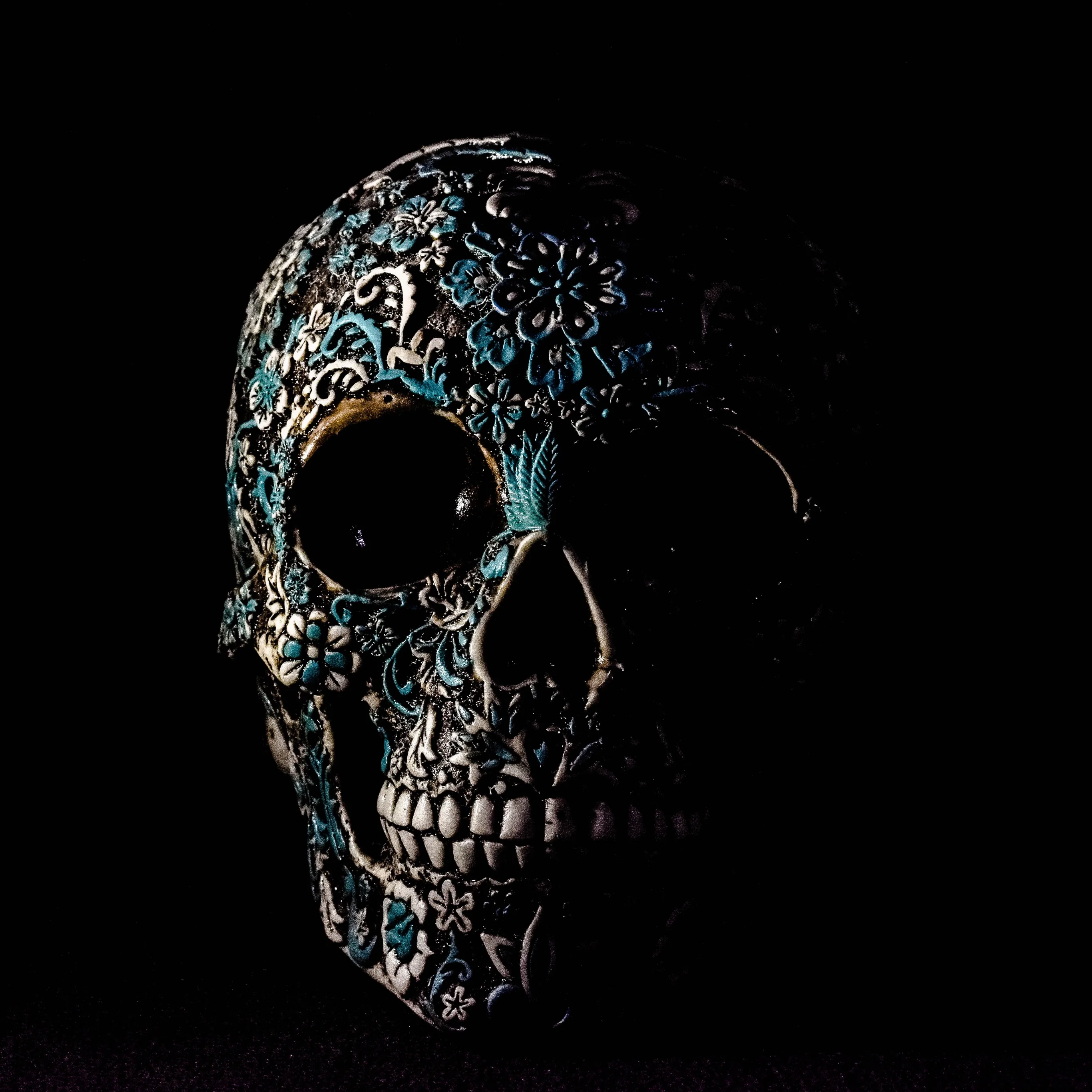 A skull covered in ornate carved filigree, turquoise inlay catching the light along the brow and cheekbone while the rest sinks into shadow.