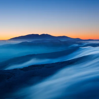 Long-exposure cloud pours like water over shadowed ridges at dawn, a thin orange horizon fading into deep blue gradients that sit well behind app icons.
