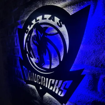A metal Dallas Mavericks logo sign glows blue against a brick wall, backlight spilling from its edges while the corners stay dark.