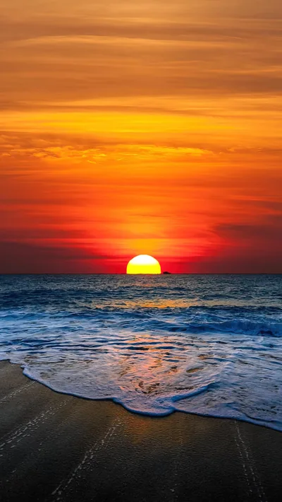 A round sun drops toward a banded orange sky while blue waves stack toward dark sand, the top third staying clear enough for a phone clock.