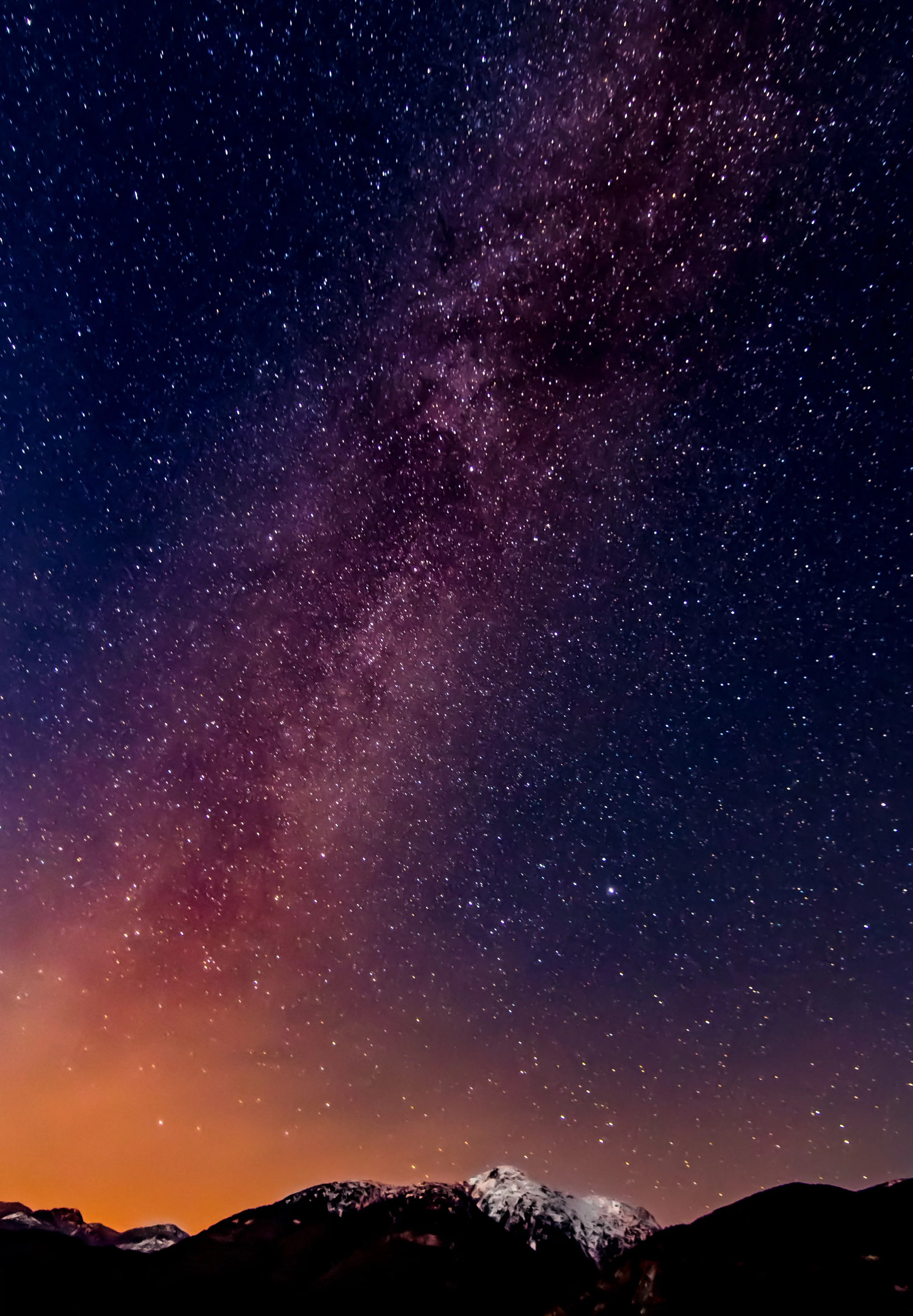 The galactic band runs diagonally through a purple sky toward a snow-capped peak, warm orange light pooling along the horizon of this portrait frame.
