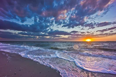 A thin sheet of foam slides up dark sand as the sun drops behind blue-grey clouds, its gold burst sitting right of centre on the horizon.