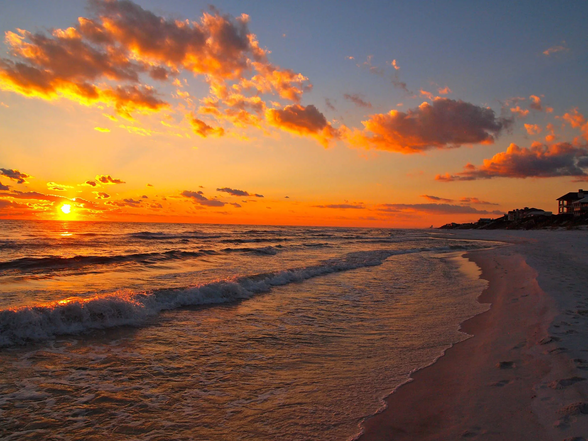 Orange-lit clouds drift over lines of surf as the sun touches the horizon on the left, beach houses standing tiny along the far right shore.