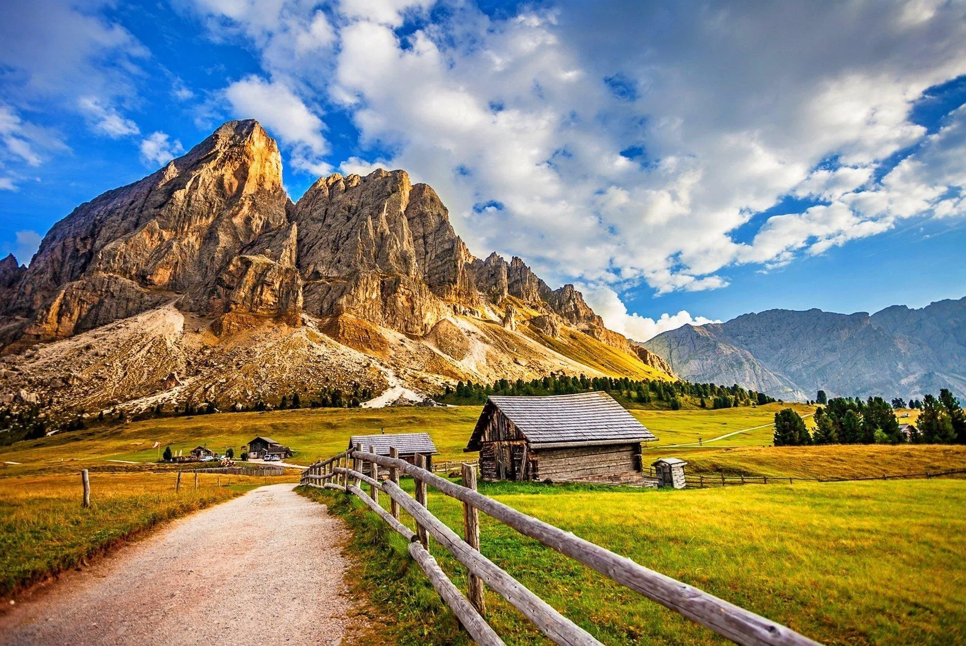 A fenced gravel lane passes a wooden barn beneath a pale rock wall, warm afternoon light on the pasture under scattered cumulus.