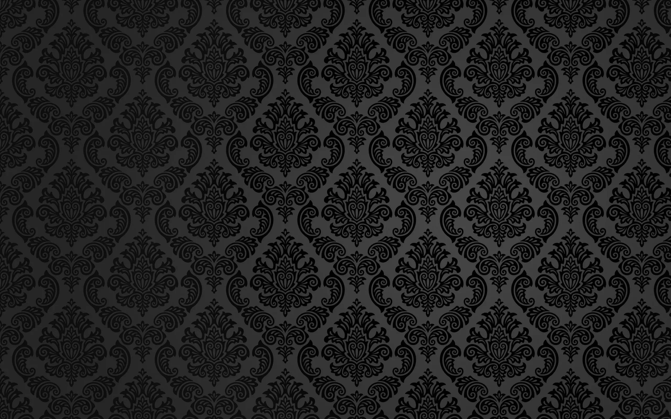 A damask repeat sits raised on charcoal, lit brightest through a horizontal band across the middle and sinking to black at the corners.
