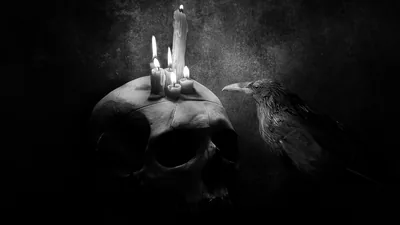 Three lit candles burn on top of a skull while a raven perches beside it, the grainy monochrome shot lit from one weak source.