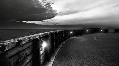 A concrete sea wall sweeps in from the left with lamps set into its face, storm cloud breaking over the water and the promenade left dark and empty.