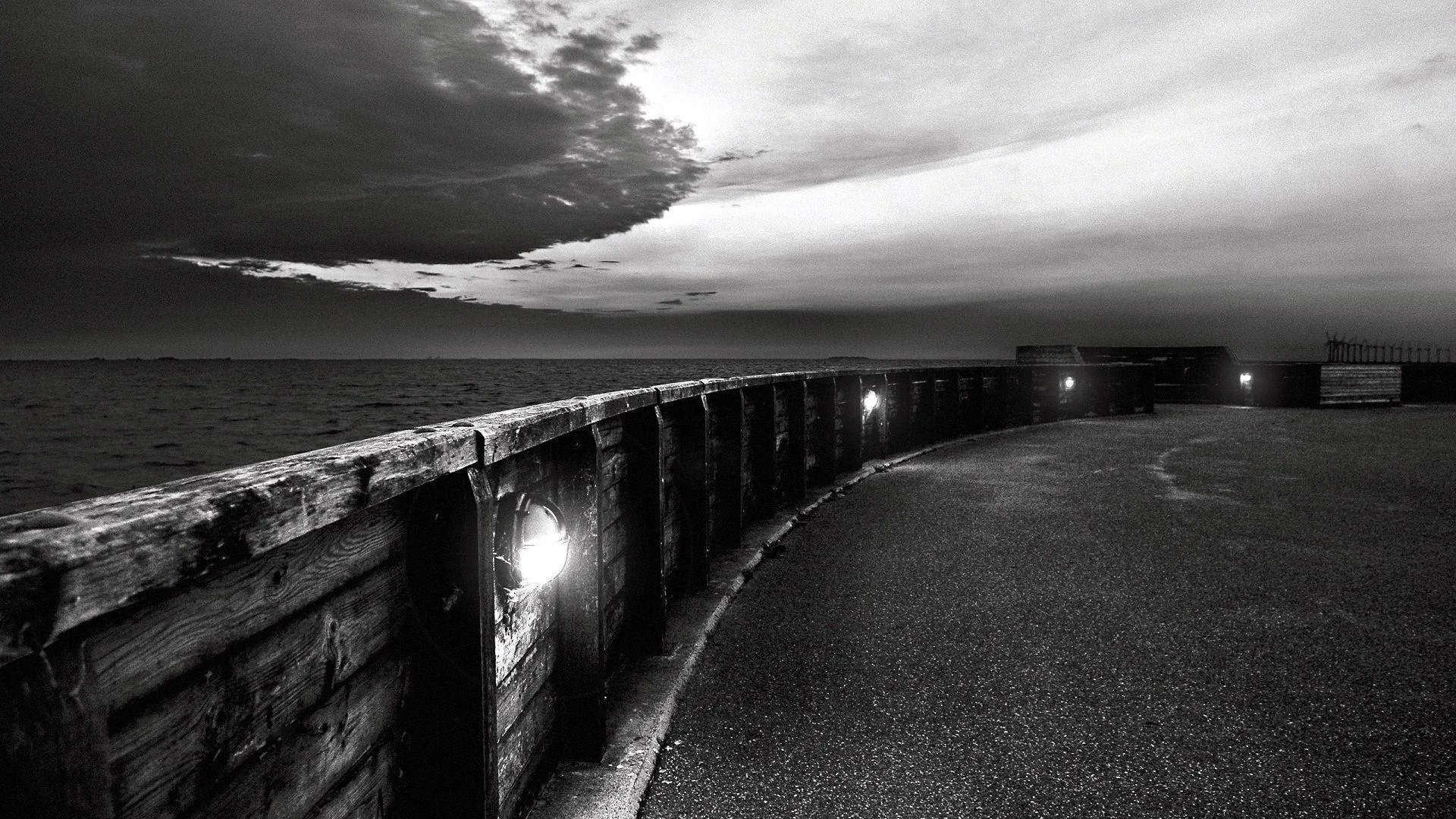 A concrete sea wall sweeps in from the left with lamps set into its face, storm cloud breaking over the water and the promenade left dark and empty.