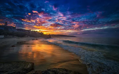 Blue and violet clouds catch a band of orange fire above a curving cove, long-exposure water smoothing the shore below a hillside town.