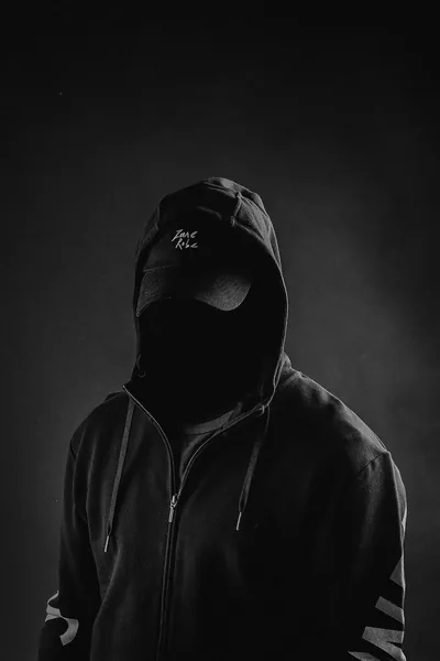 A figure in a zipped black hoodie and balaclava stands in soft studio haze, monochrome with the face left as a flat dark oval.