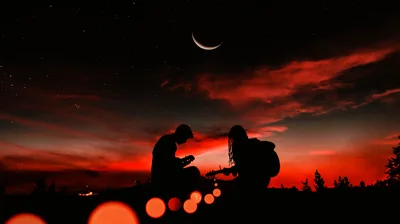 Two people crouch close together on a hilltop while a crescent moon hangs in banded crimson cloud, out-of-focus orange lights blurring across the bottom of the frame.