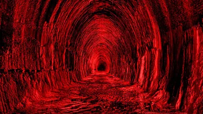 A long stone tunnel recedes to a black vanishing point, the rough walls and rail bed washed in a single red light.