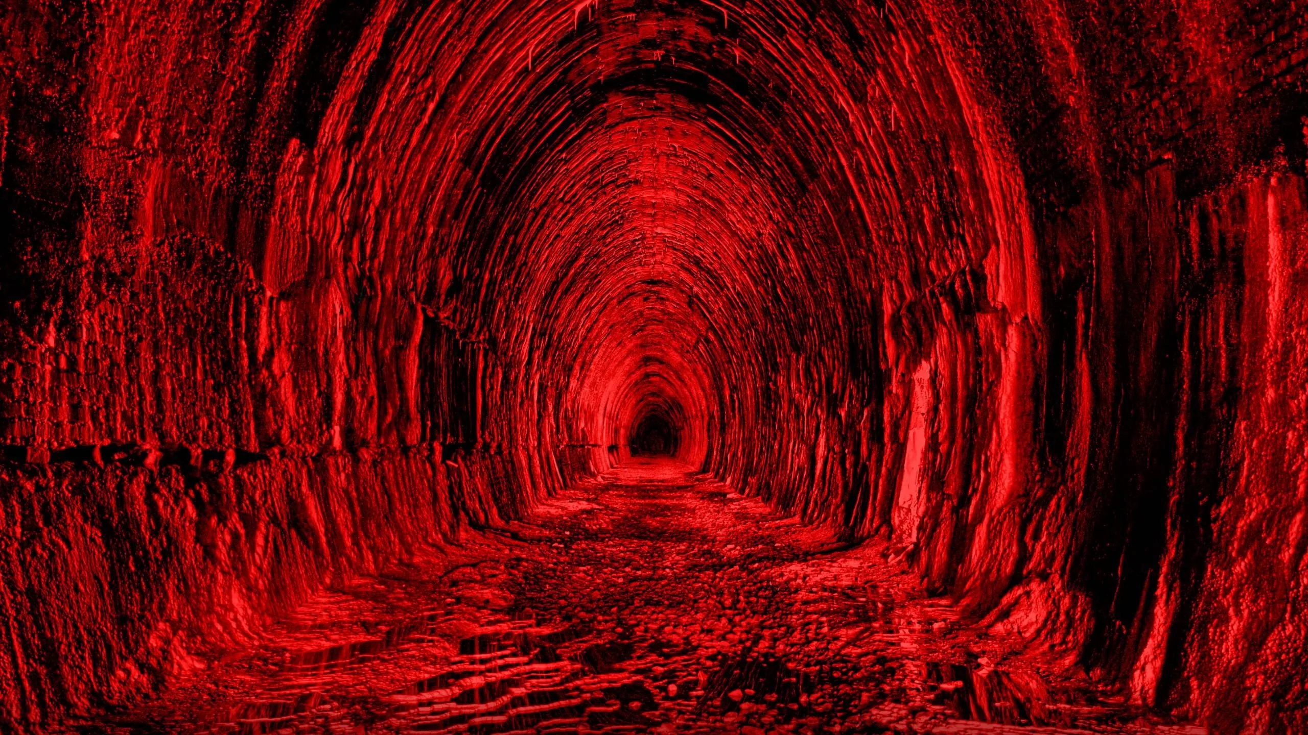 A long stone tunnel recedes to a black vanishing point, the rough walls and rail bed washed in a single red light.