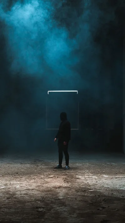 Teal smoke hangs in the rafters above a man facing a dimly lit doorframe, the sandy floor and black side walls leaving both edges free for icons.