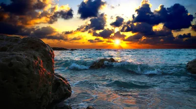 The sun flares between heavy clouds and lights the tops of wet boulders in the foreground, teal waves churning through the rocks below.