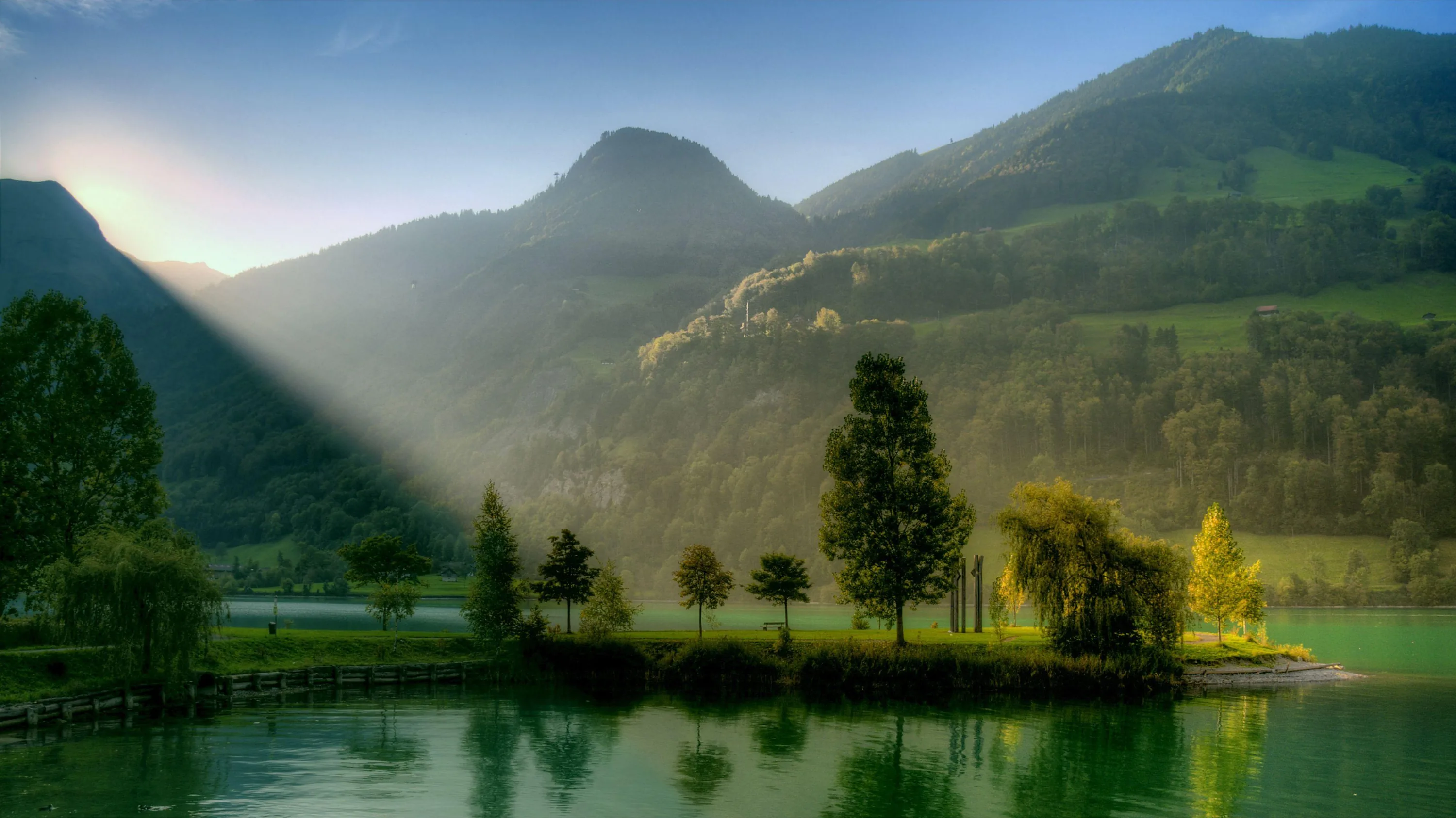 A single shaft of sun breaks over the ridge at top left, spotlighting a tree-lined point while haze mutes the mountains to soft grey-green.