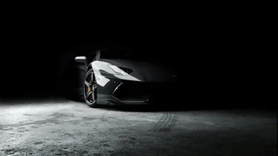 One overhead light picks out the roof, wing mirror and front splitter of an Aventador on a rough concrete floor, everything past the car left black.