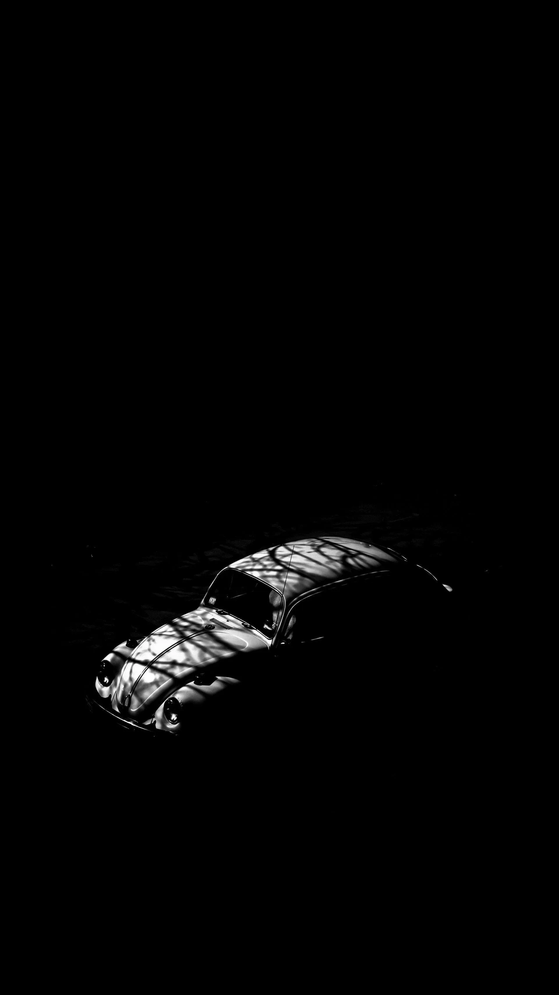 A rounded vintage car sits in near-total darkness, one overhead light picking out the roof and one wheel arch.