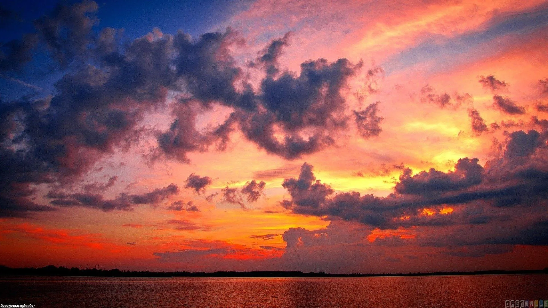 Broken dark clouds hang over a still lake at sundown, their orange-lit edges doubled in the water along the bottom of the frame.