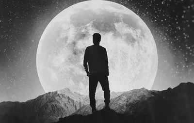 Shot in monochrome, a man stands on a ridge dwarfed by the full moon filling most of the frame, stars speckling the black sky at right.