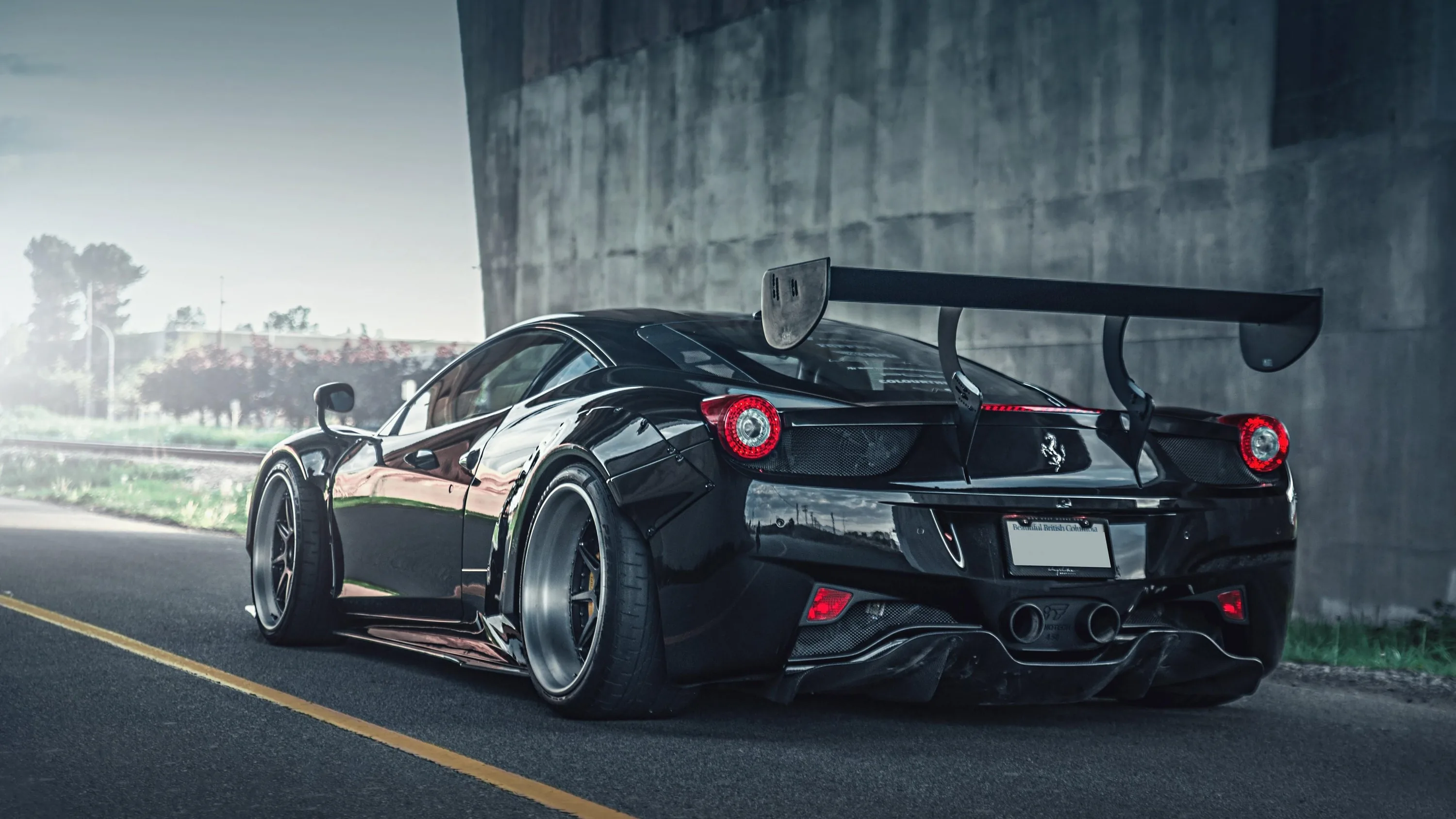Rear three-quarter of a track-prepped 458 under a concrete underpass, its swan-neck wing and wide fenders framed by roadside grass and daylight beyond.