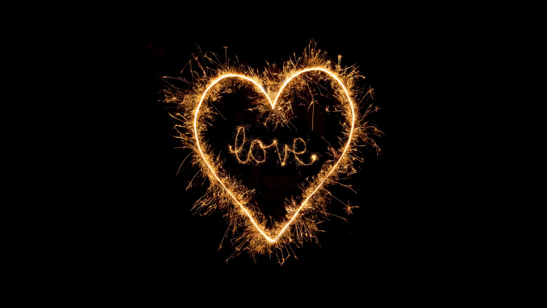 A heart drawn in burning sparkler trails on pure black, the word love written in the same gold script at its centre.