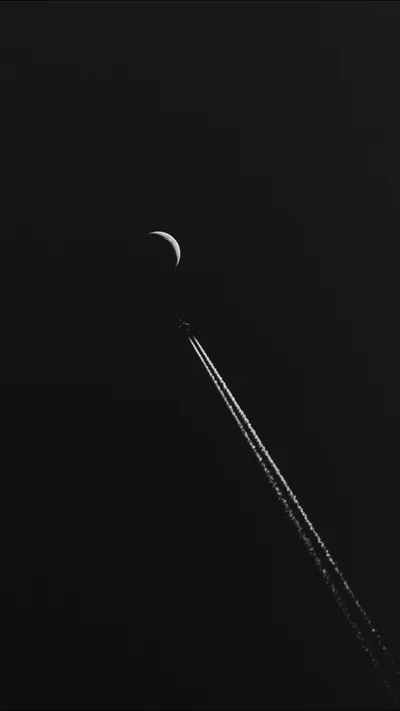 A jet contrail cuts a thin diagonal line below a barely lit crescent, the rest of the tall frame sunk in near-total black.