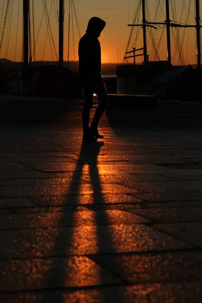 A boy walks across wet dock stones as the sun drops behind moored masts, his long shadow cut into the orange reflection.