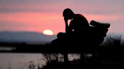 Head resting on one hand, a man sits on a park bench while the sun melts into the horizon behind him, pink haze filling the upper right.