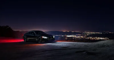 Shot from a gravel ridge, the car's headlights point out over a valley of orange city lights, red light painting spilling across the ground behind the rear wheels.