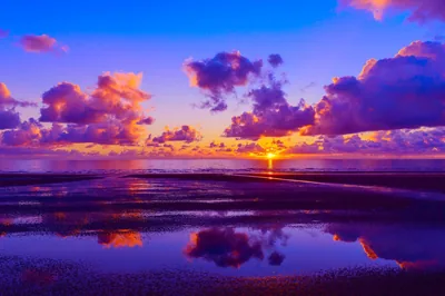 Puffy clouds glow pink and violet around a low sun, repeating in sheets of water across the beach, the saturation pushed far past natural.