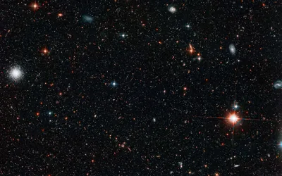 A deep field crowded with tiny galaxies and pinprick stars, one white star burning at the left edge and a red-orange one low on the right.