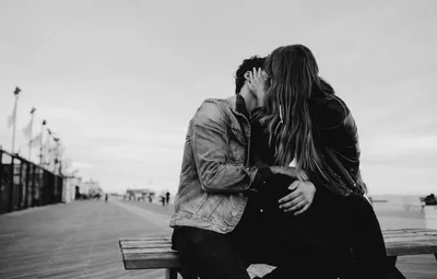 Two people lean into each other on a bench at the end of a wooden pier, the sky behind them blown out to near white.