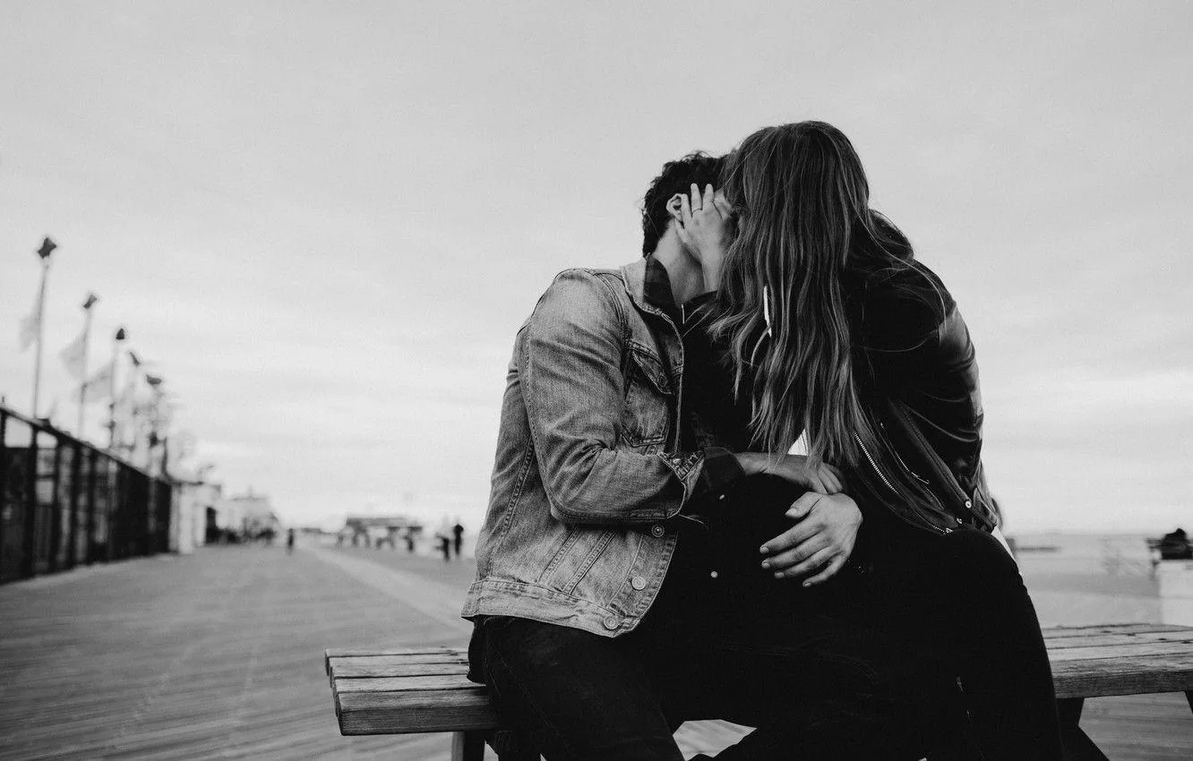Two people lean into each other on a bench at the end of a wooden pier, the sky behind them blown out to near white.