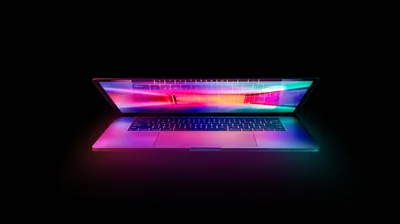 A half-open laptop throws pink and blue light onto a black surface, the screen edge glowing along its length.