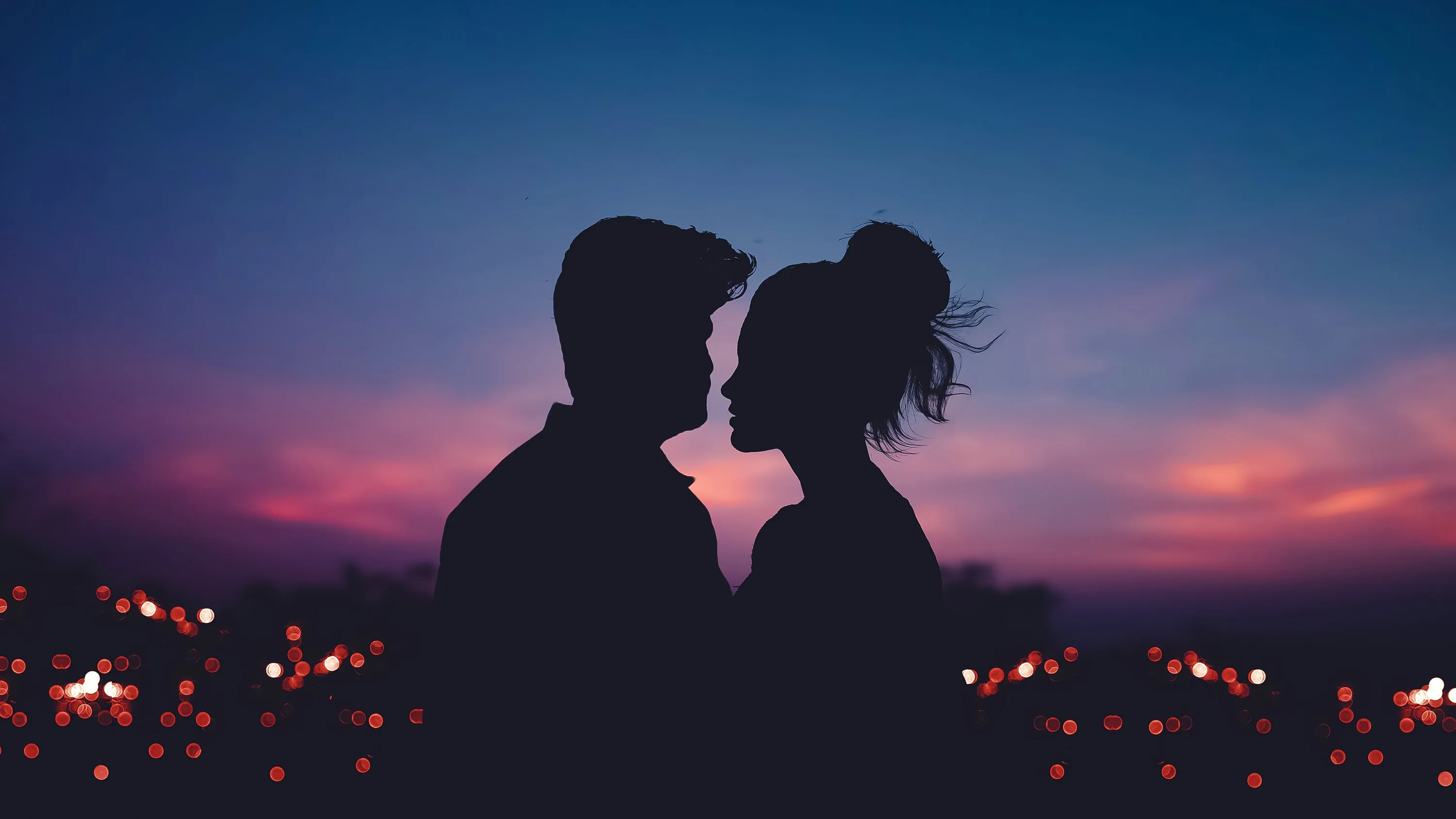 A couple stand nose to nose as the sky fades from indigo to pink behind them, blurred amber lights scattered along the bottom third of the frame.