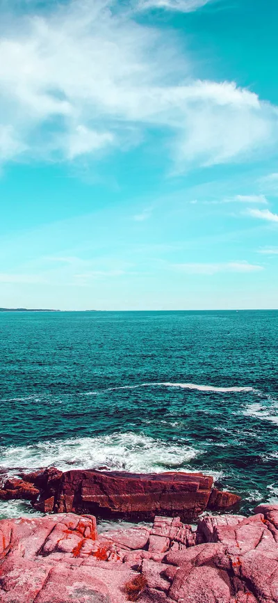 Reddish-pink boulders edge a teal sea under a pale filtered sky, the top half left mostly empty for a phone clock.