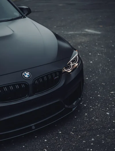 The nose of a matte black BMW shot close up on wet asphalt, only the headlight ring and the roundel catching any light.