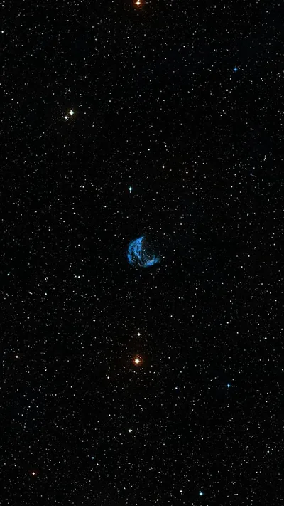 A small blue crescent of gas sits alone at the centre of a wide black field, orange and white stars scattered far around it.