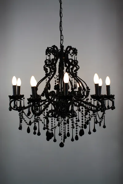 A black baroque chandelier hung with dark glass droplets, its white candle bulbs lit against a plain pale-grey wall that fills every corner.
