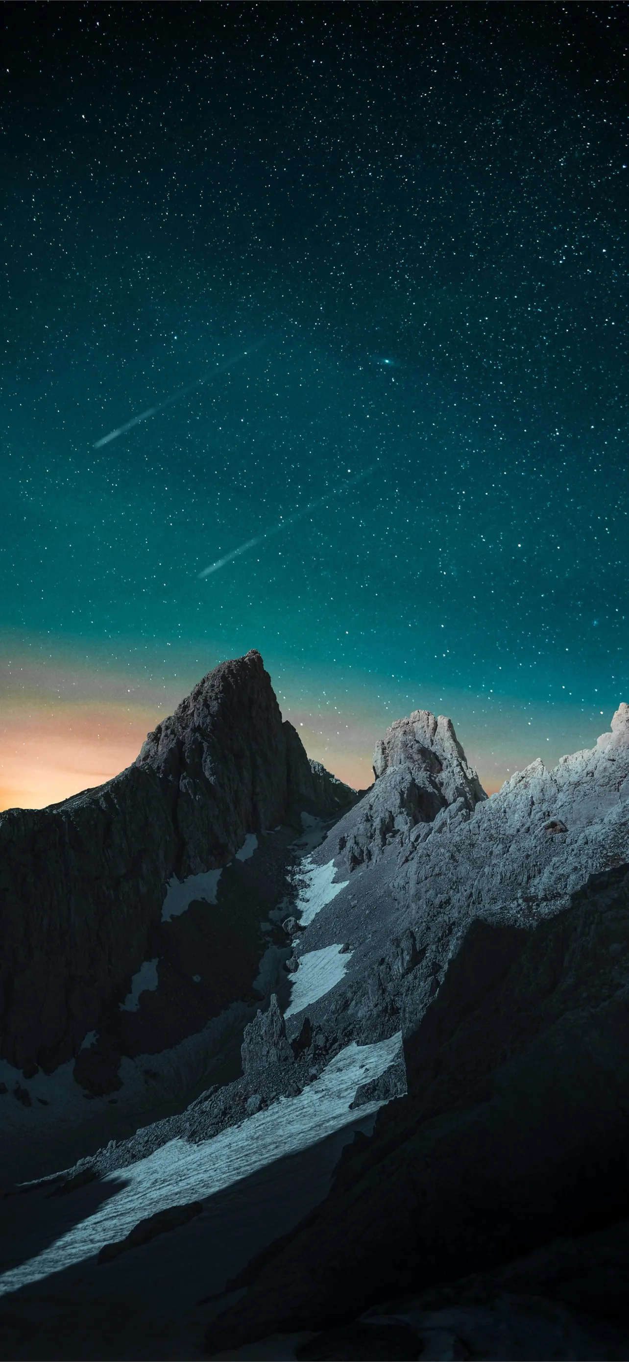 Two faint meteor streaks cross the starry sky above a jagged black summit, a low orange glow at the horizon and snow catching pale light lower right.