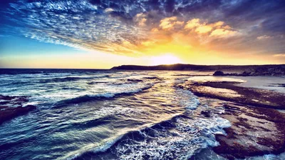 Dappled clouds catch first light above surf threading through flat coastal rock, distant hills holding the horizon line just below centre.
